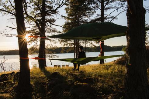 Person sitter i ett dragtält uppställt mellan träd. Han tittar på vattnet som syns framför honom. Solen går ner och färgar landskapet.