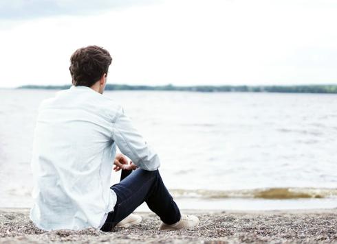 Takaa kuvattu aikuinen istuu yksin rantahiekalla ja katselee ulapalle.