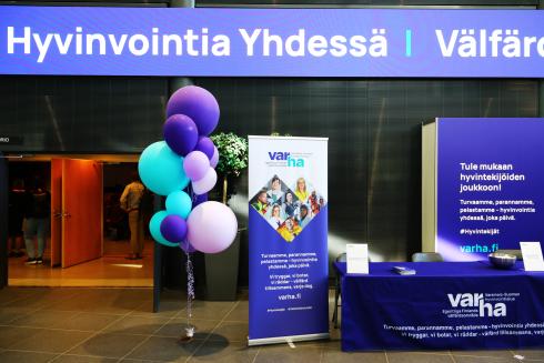 On the left, purple and turquoise balloons on a stick. On the right side of the balloons, Varha's roll up and the fair counter. Above them, a light board with the words "Hyvinvointia yhdessä" (in English "Well-being together").