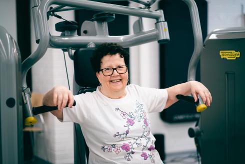 En äldre dam sitter i en maskin på gymmet och ler glatt mot kameran.