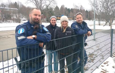 Kaksi poliisia ja kaksi muuta henkilöä nojaavat verkkoaitaan ja hymyilevät. 