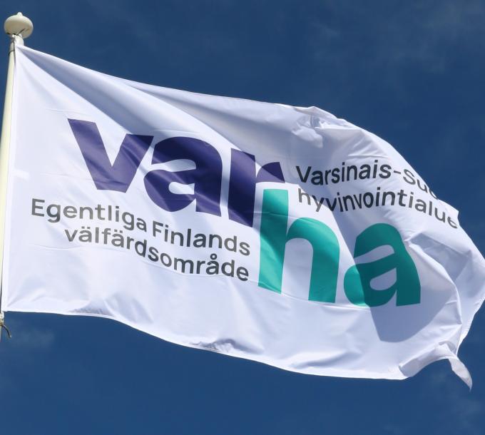 Valkoinen lippu lipputangossa. Lipussa on logo, jossa lukee ”Varha. Varsinais-Suomen hyvinvointialue. Egentliga-Finlands välfärdsområde”. Taivas taustalla on kirkkaan sininen.