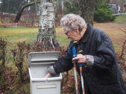 Harmaahiuksinen nainen ottaa toseilla kädellään postilaatikosta posteja. Toisessa kädessä hänellä on kävelysauvat.